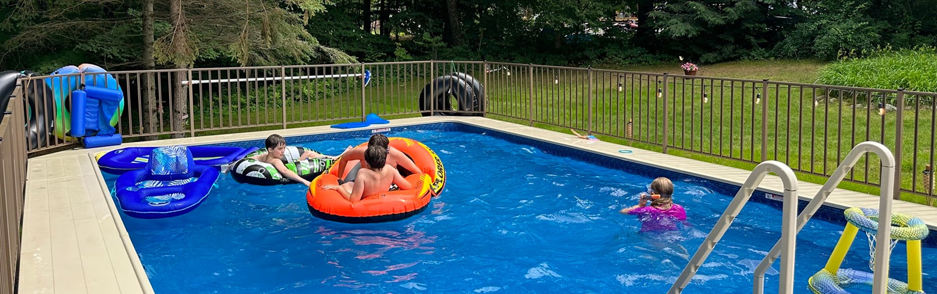 A bright suburban backyard with a swimming pool, patio seating, and a white house under clear skies.