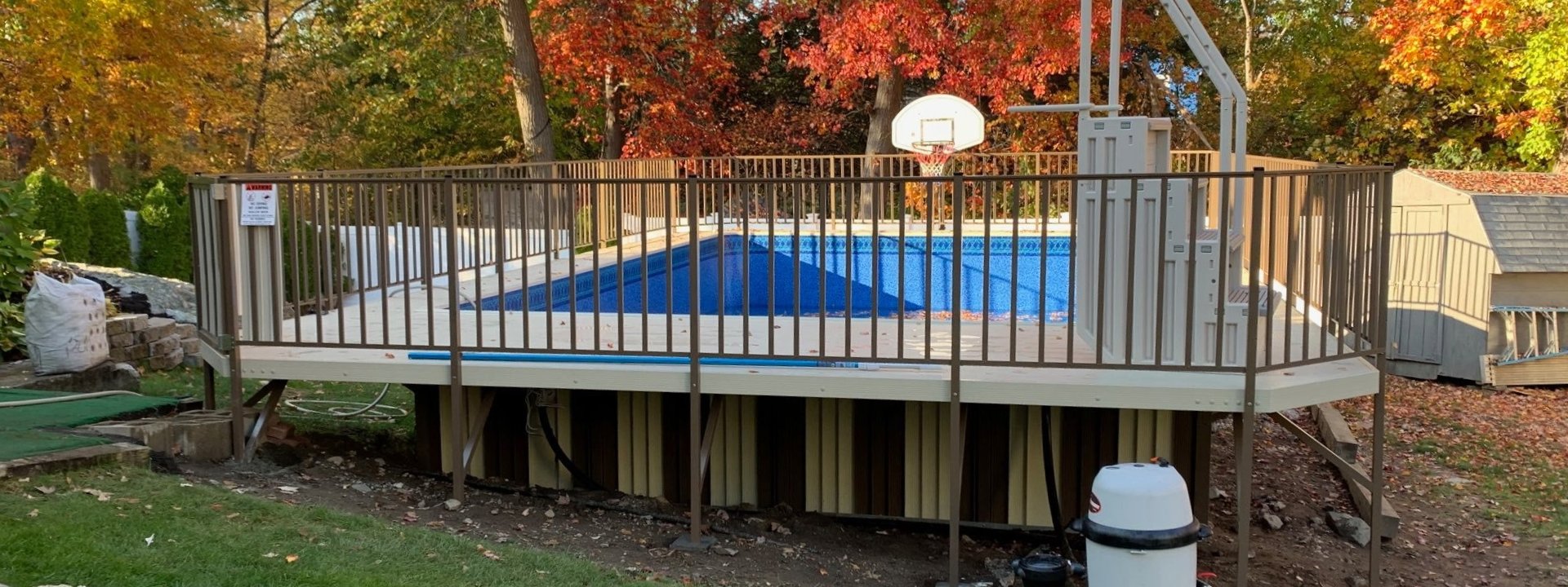 A bright suburban backyard with a swimming pool, patio seating, and a white house under clear skies.