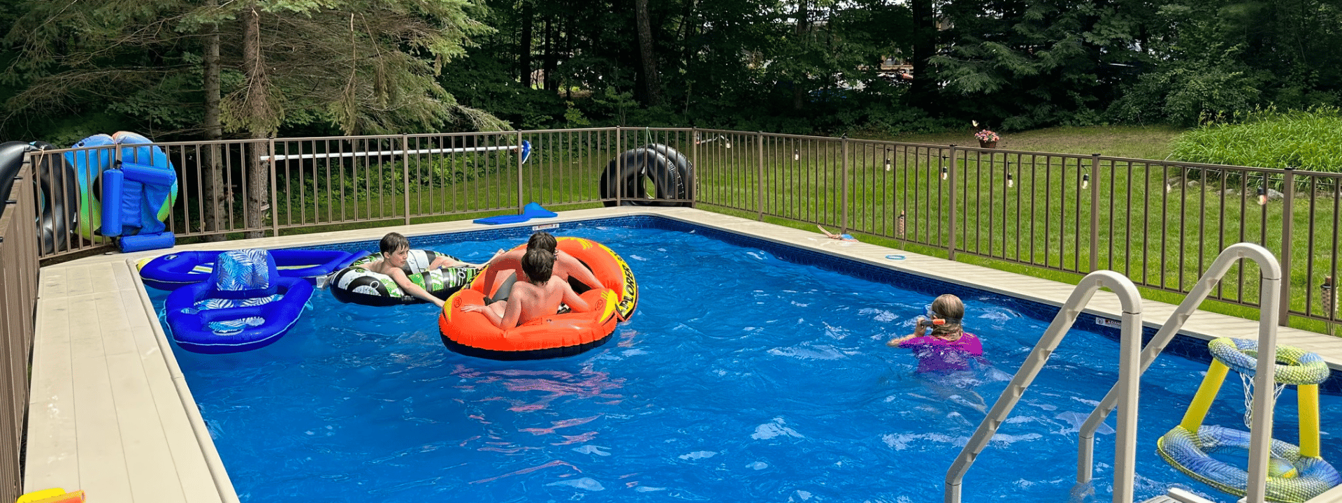 A girl in a colorful swimsuit steps out of a backyard swimming pool while a boy rests on the pool edge, with two adults sitting nearby at a large suburban home.